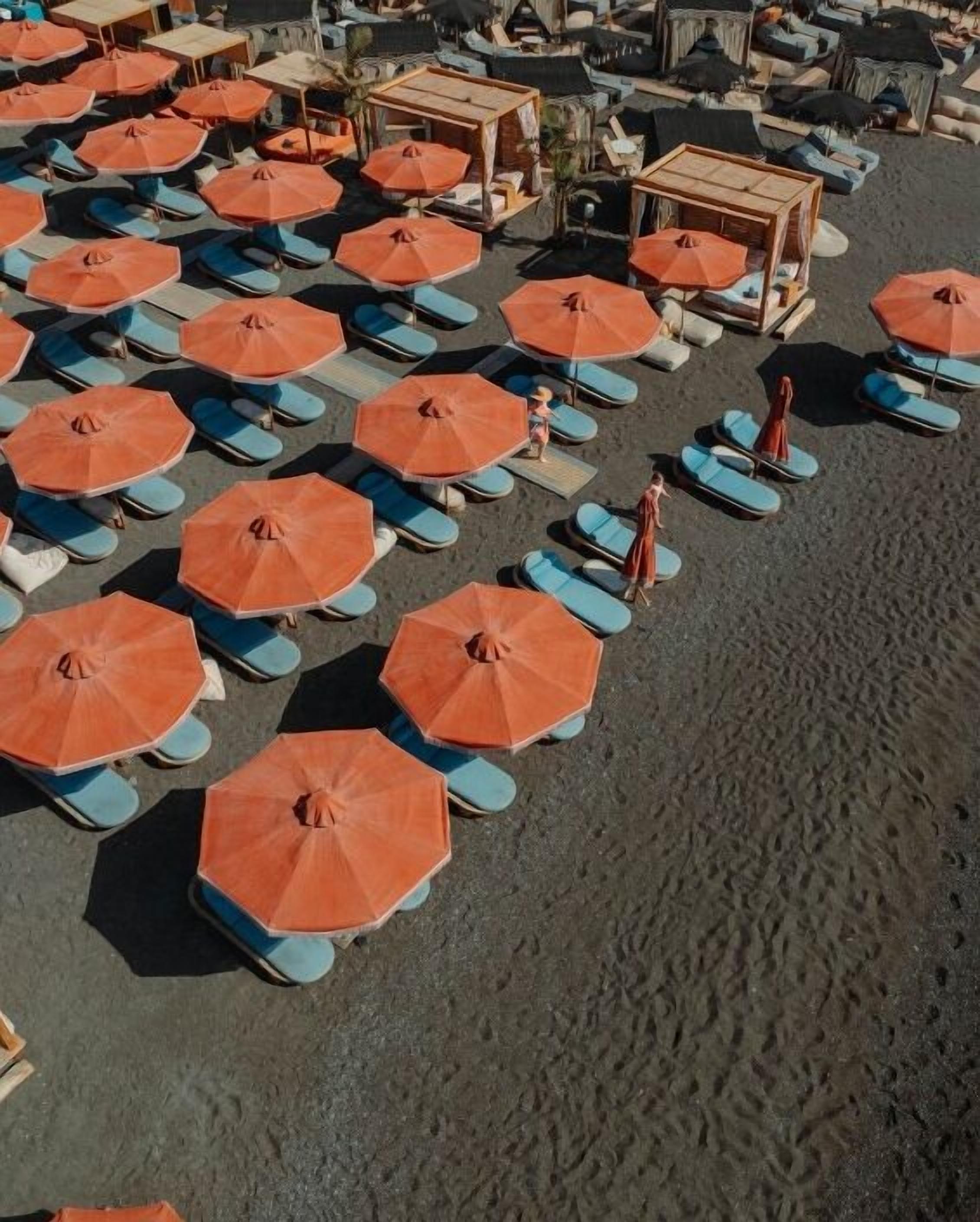 An aerial view of a beach with rows of sun loungers and orange umbrellas.