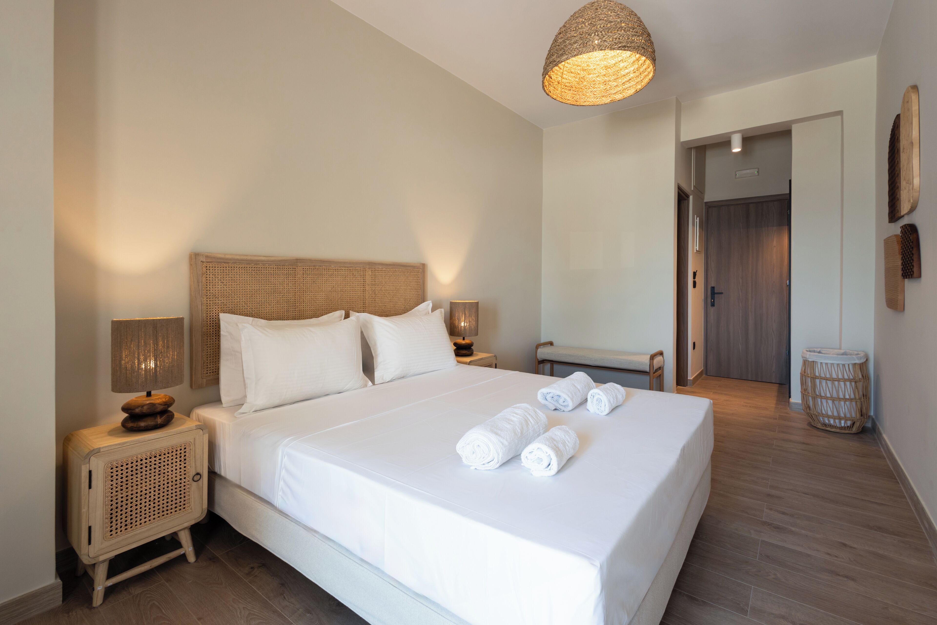 A neatly made bed with white linens and rolled towels, a wicker headboard, and a bedside lamp on a wooden nightstand.