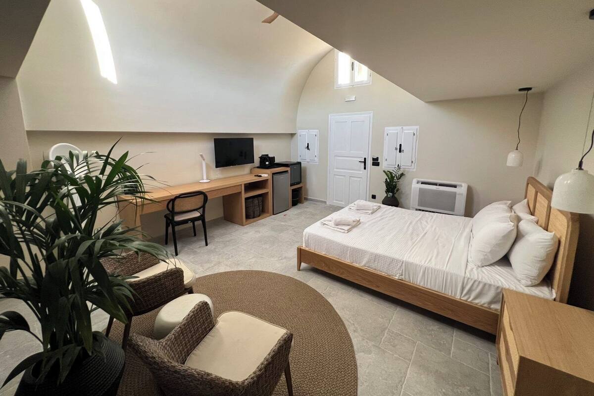 A compact, modern bedroom with a loft-style ceiling, a bed, a desk, a chair, and a small seating area.