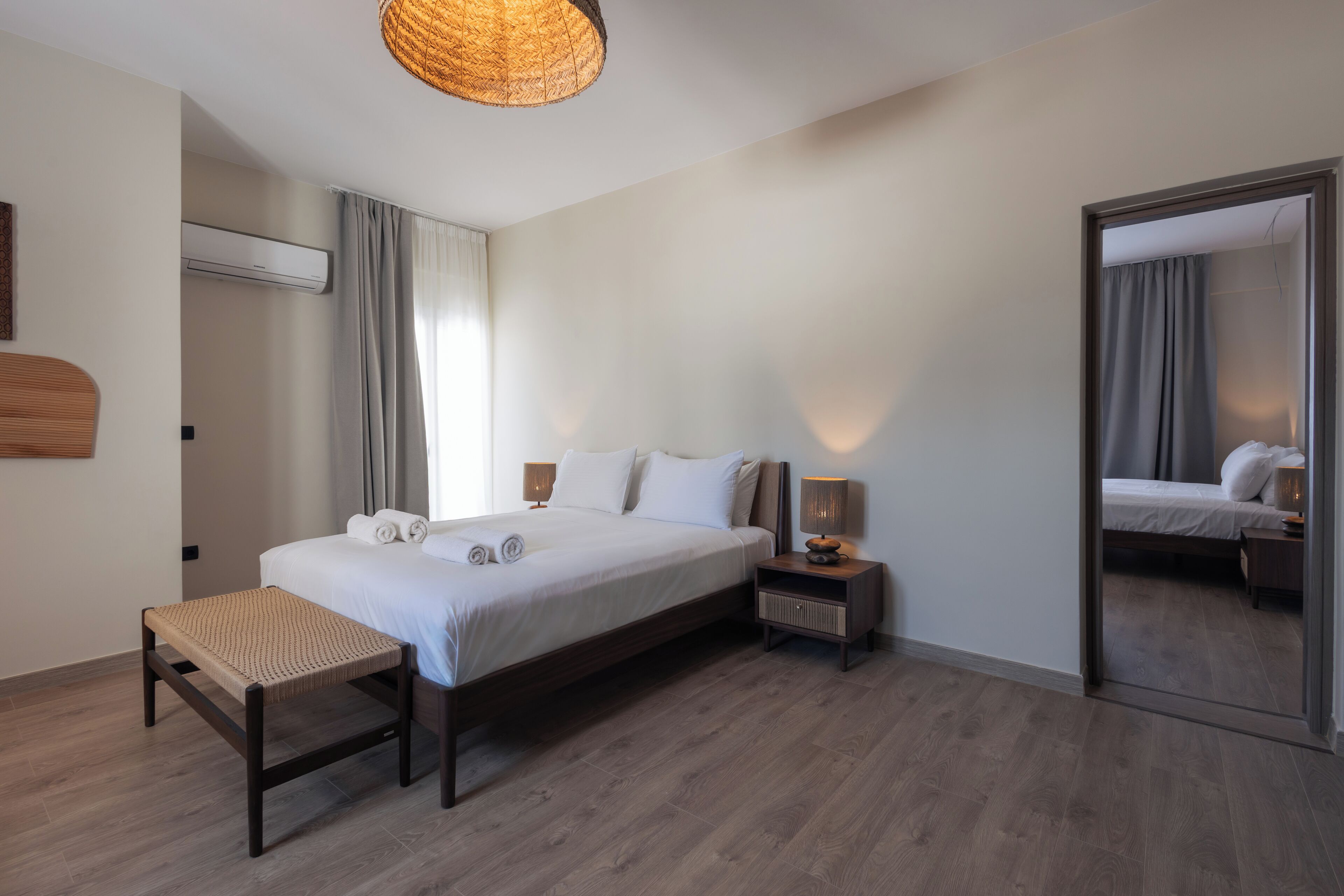 A modern hotel room with a large bed, a small bedside table, a bedside lamp, a woven stool, and a mirror on the wall.