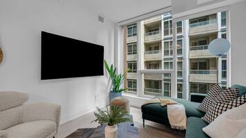 A modern living room with a large TV, a green sofa, a beige armchair, and a potted plant.