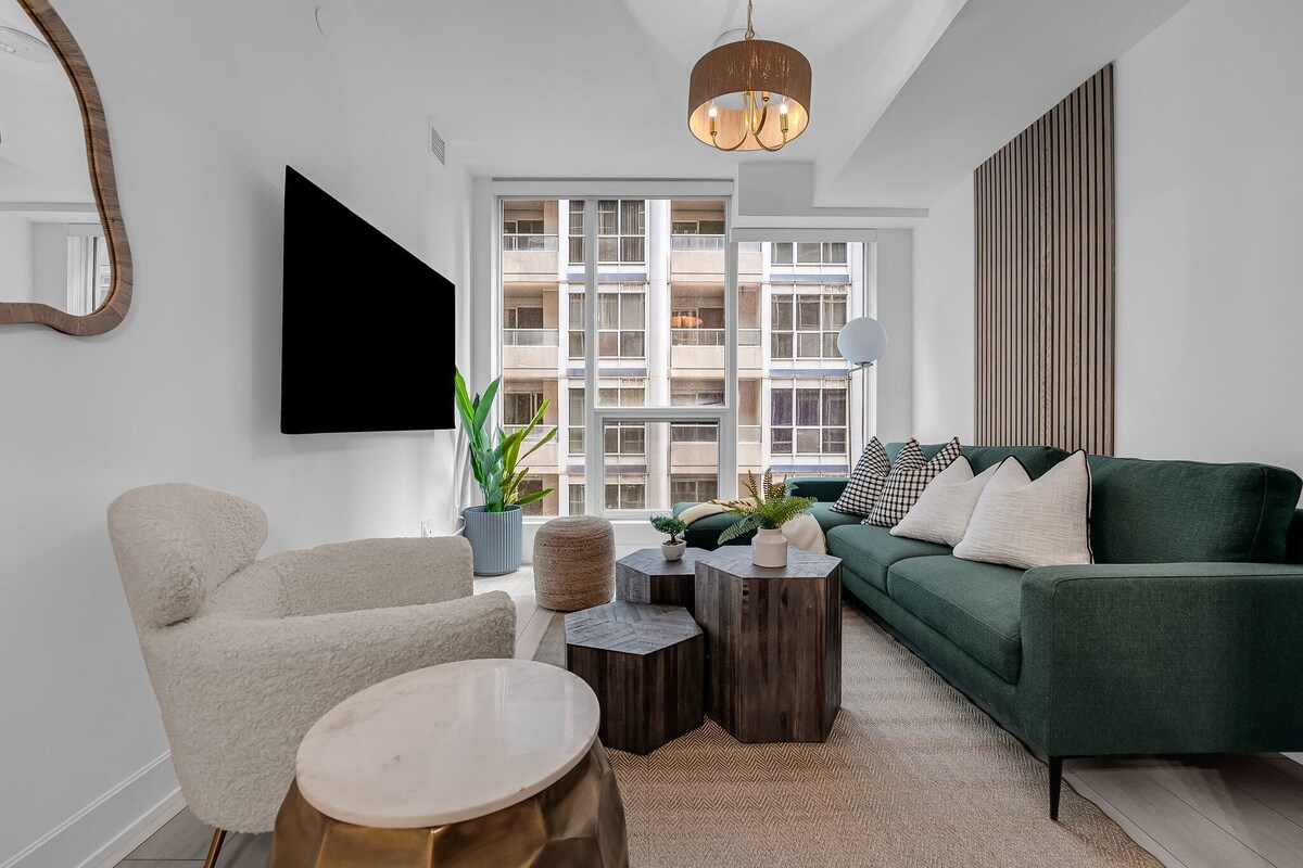 Una sala de estar moderna con un sofá verde, un sillón blanco y una mesa de centro redonda. Hay un ventanal con vistas a edificios, una planta en maceta y un televisor de pantalla plana montado en la pared.