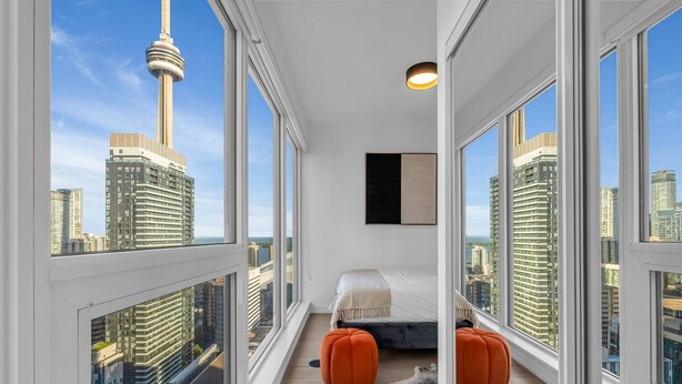 A modern bedroom with a bed, two orange ottomans, a black artwork, and large windows offering a city skyline view. - The Aurelien Luxe Collection (Toronto)