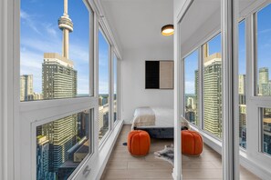Un dormitorio moderno con una cama, dos otomanas naranjas, una obra de arte negra y ventanas amplias con vistas al horizonte de la ciudad.