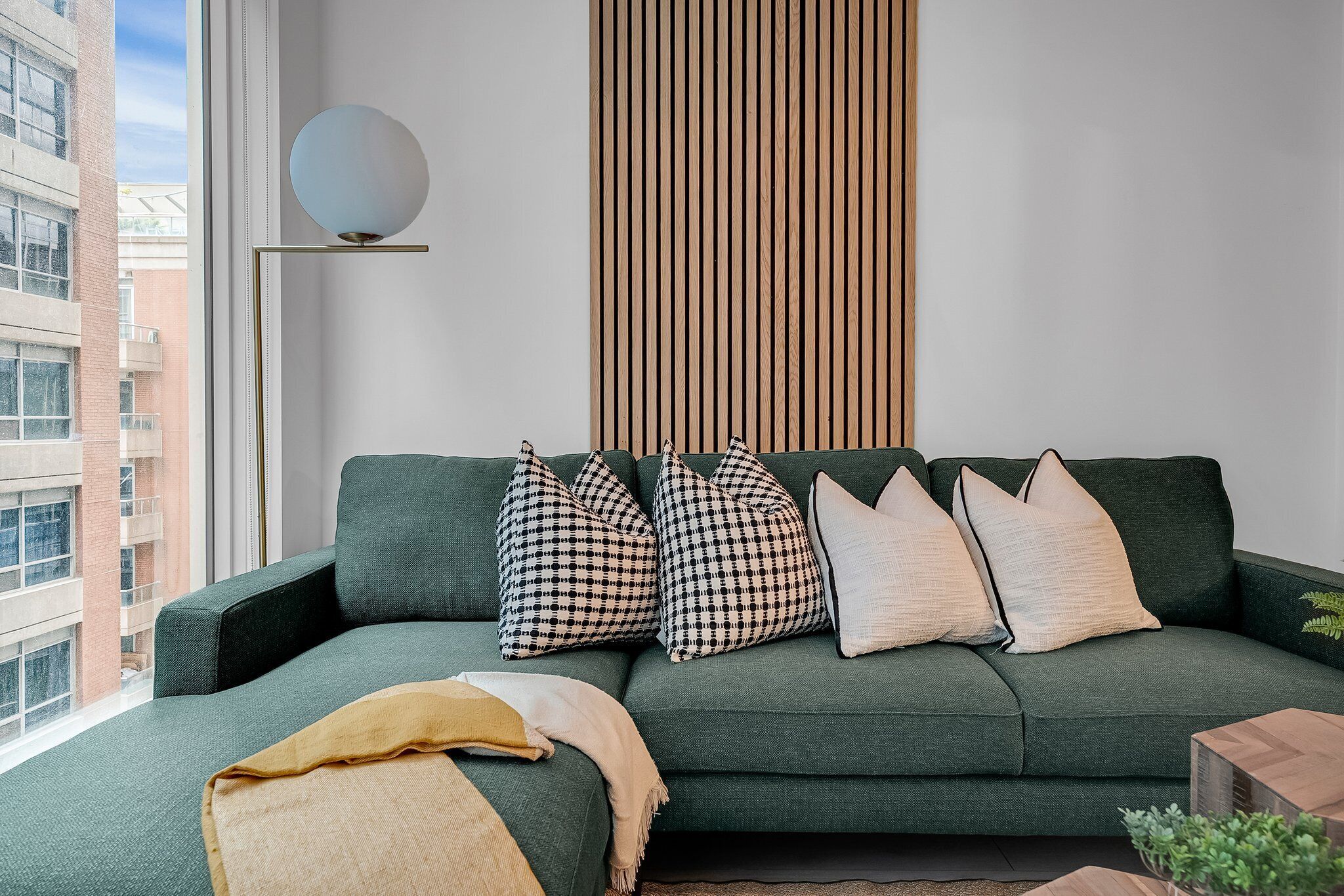 Un salón moderno con un sofá verde, cojines a cuadros, una lámpara redonda fijada a la pared y un panel de madera vertical.