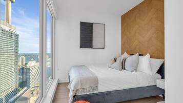 Quarto moderno com uma grande janela, cama com roupa de cama branca, cabeceira de madeira e vista para a cidade.
