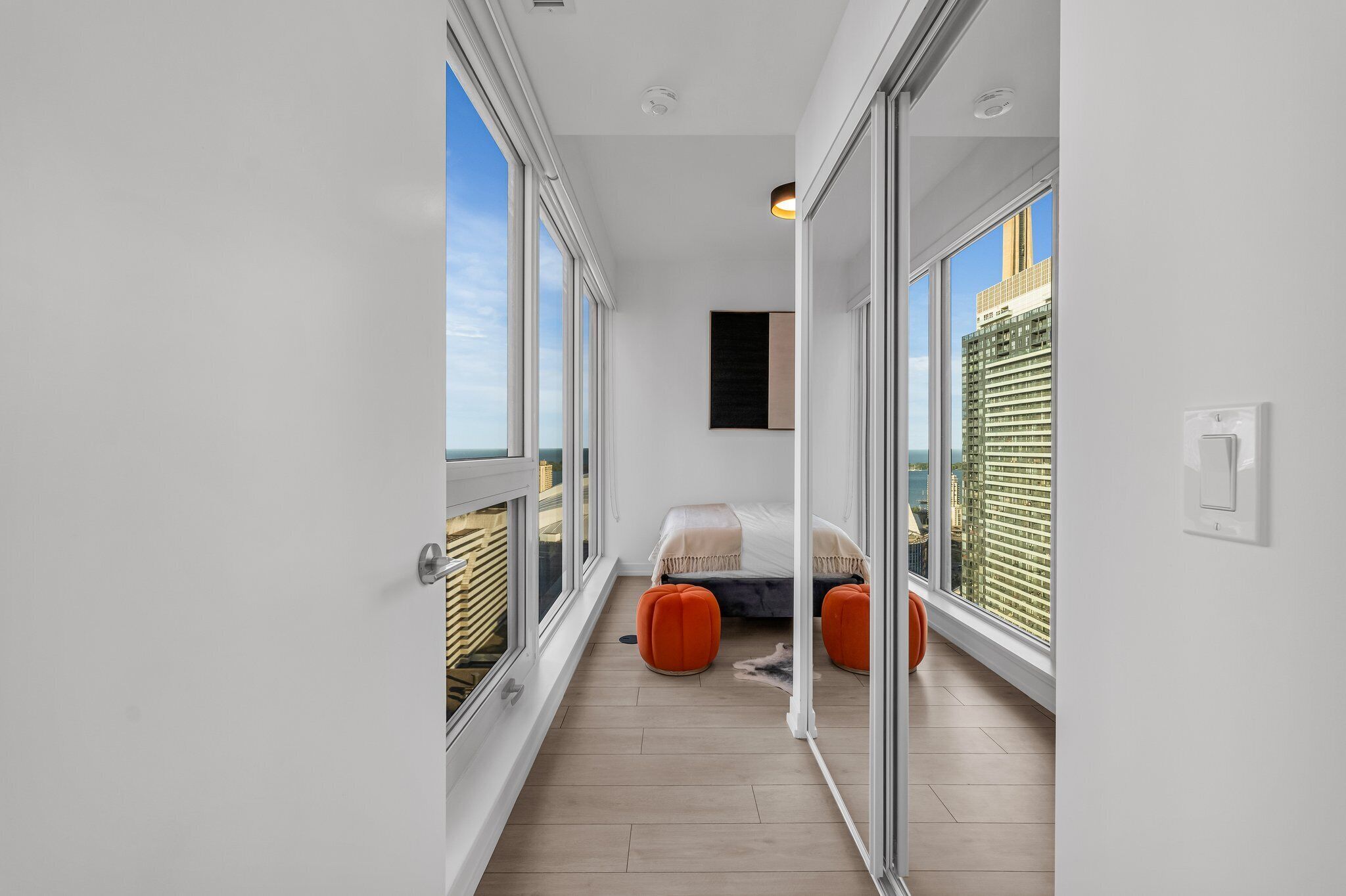A narrow balcony with a bed and orange ottomans, large windows, and a city view.