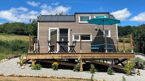 A two-story wooden cabin with a deck, outdoor seating, and a large umbrella. - Tiny House La Samorienne, with private spa and sauna. (Réjaumont)