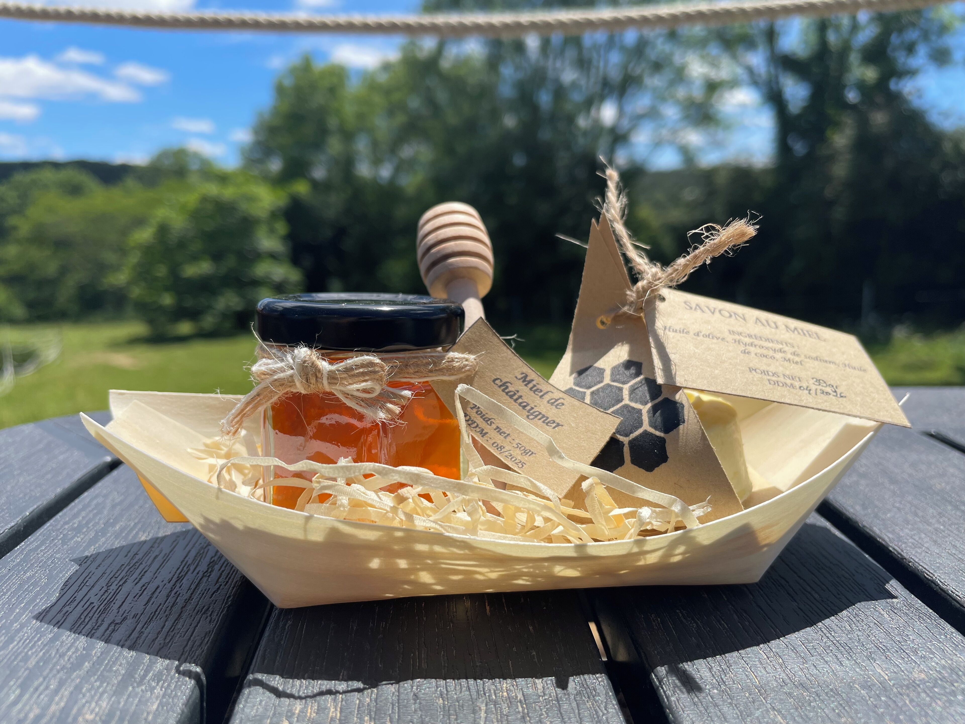 Un pot de miel doté d’une anse en bois et d’une étiquette, placé dans un panier en papier sur une surface en bois.