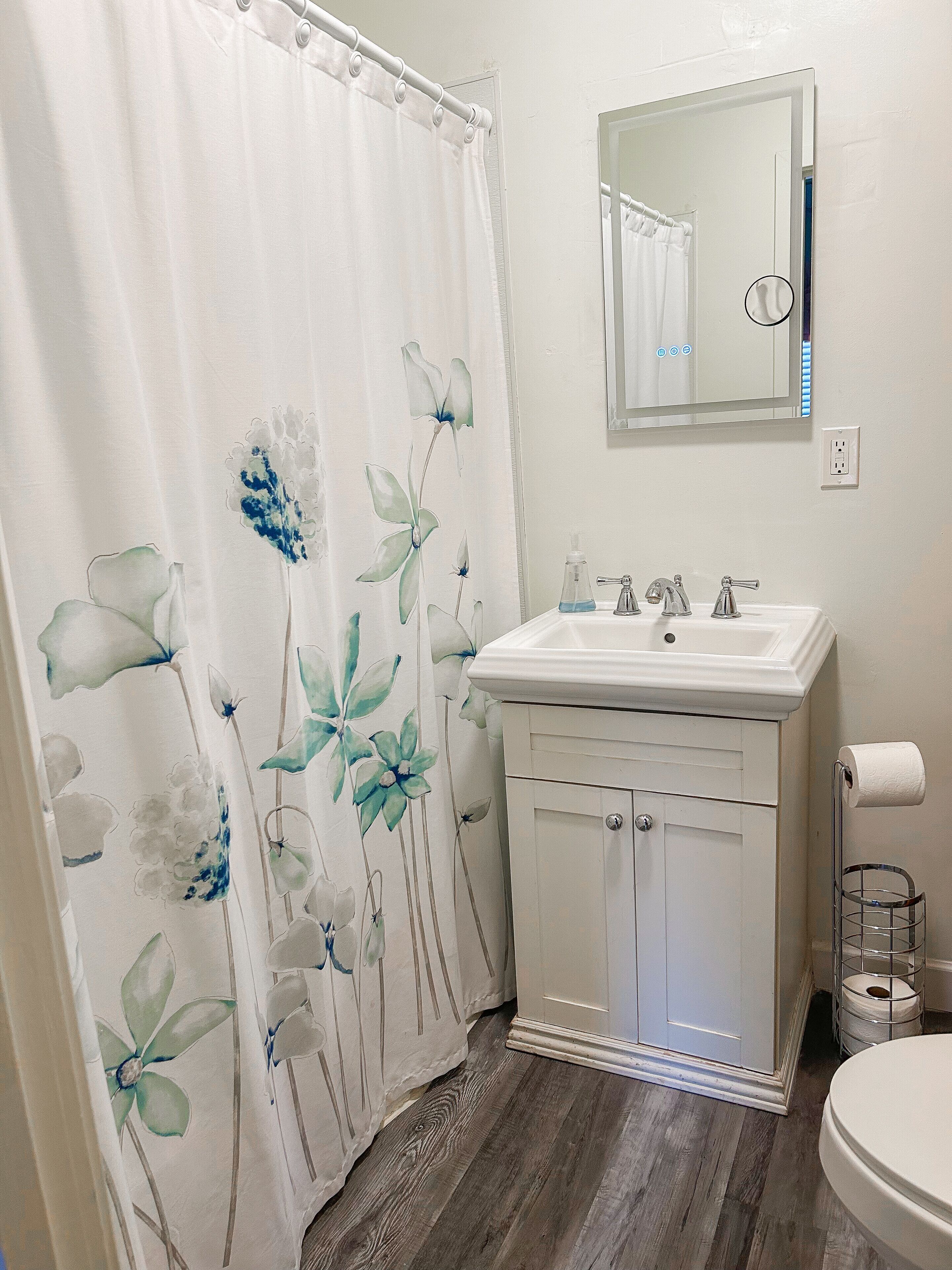 Un baño con cortina de ducha con motivos florales, un lavabo blanco con espejo arriba y un inodoro con dispensador de toallas de papel.