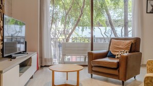 A living room with a brown leather sofa, a small wooden coffee table, a TV on a white stand, and a large window with a view of trees and a balcony. - New Apartment City Oasis In Roma Norte (Mexico City)