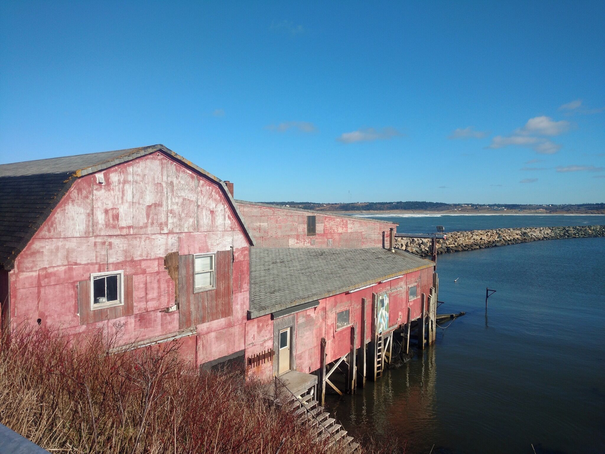 Ein rotes Holzgebäude mit geneigtem Dach, gelegen an einem ruhigen Gewässer, vermutlich ein Hafen oder eine Bucht, mit einem steinernen Wellenbrecher im Hintergrund.