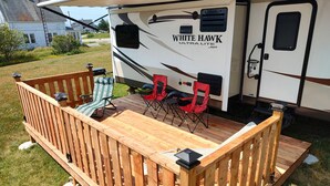 Een recreatiewagen met een houten terras, twee opklapbare stoelen en een barbecuehoek.