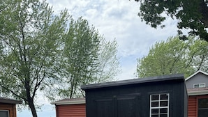 Un petit abri noir avec une fenêtre, entouré d’une cour verdoyante et bordé d’arbres en arrière-plan.