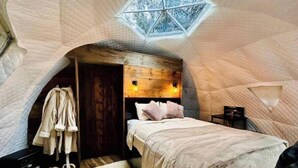 A cozy bedroom with a wooden headboard, a bed with pillows, a hanging robe, and a small table with a lamp.