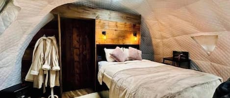 Um quarto aconchegante com cabeceira de madeira, cama com almofadas, roupão pendurado e uma pequena mesa com abajur.