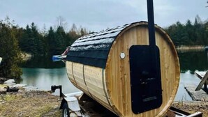 A wooden barrel-shaped structure with a chimney, positioned by a calm body of water with a forested background.