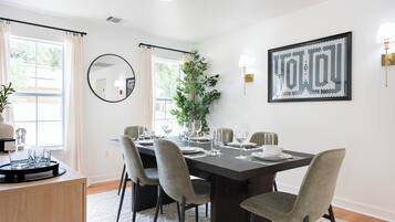 Uma sala de jantar com uma mesa de madeira escura posta para quatro pessoas, rodeada por cadeiras estofadas cinzentas. Um espelho grande está pendurado na parede, e uma obra de arte abstrata emoldurada é exibida acima da mesa.