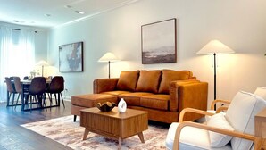 Una sala de estar moderna con un sofá de cuero marrón, una mesa de centro de madera y un sillón blanco. En el fondo, se observa un comedor con una mesa y sillas.