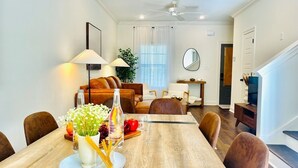 Un comedor con una mesa de madera, sillas y un florero con flores. Se observa un sofá y un televisor al fondo.