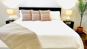 Una cama bien hecha con edredón blanco, varios almohadones y una manta color beige. Hay dos mesitas de noche con lámparas y una planta en maceta.