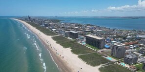Vista aérea de uma cidade costeira com uma extensa praia, vários edifícios altos e uma ponte ao longe.