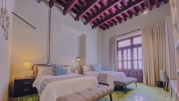 A bedroom with two beds, a bench, a nightstand, a lamp, and a window with curtains. - Beautiful double suite in an old house with contemporary restoration.  (Heroica Puebla de Zaragoza)
