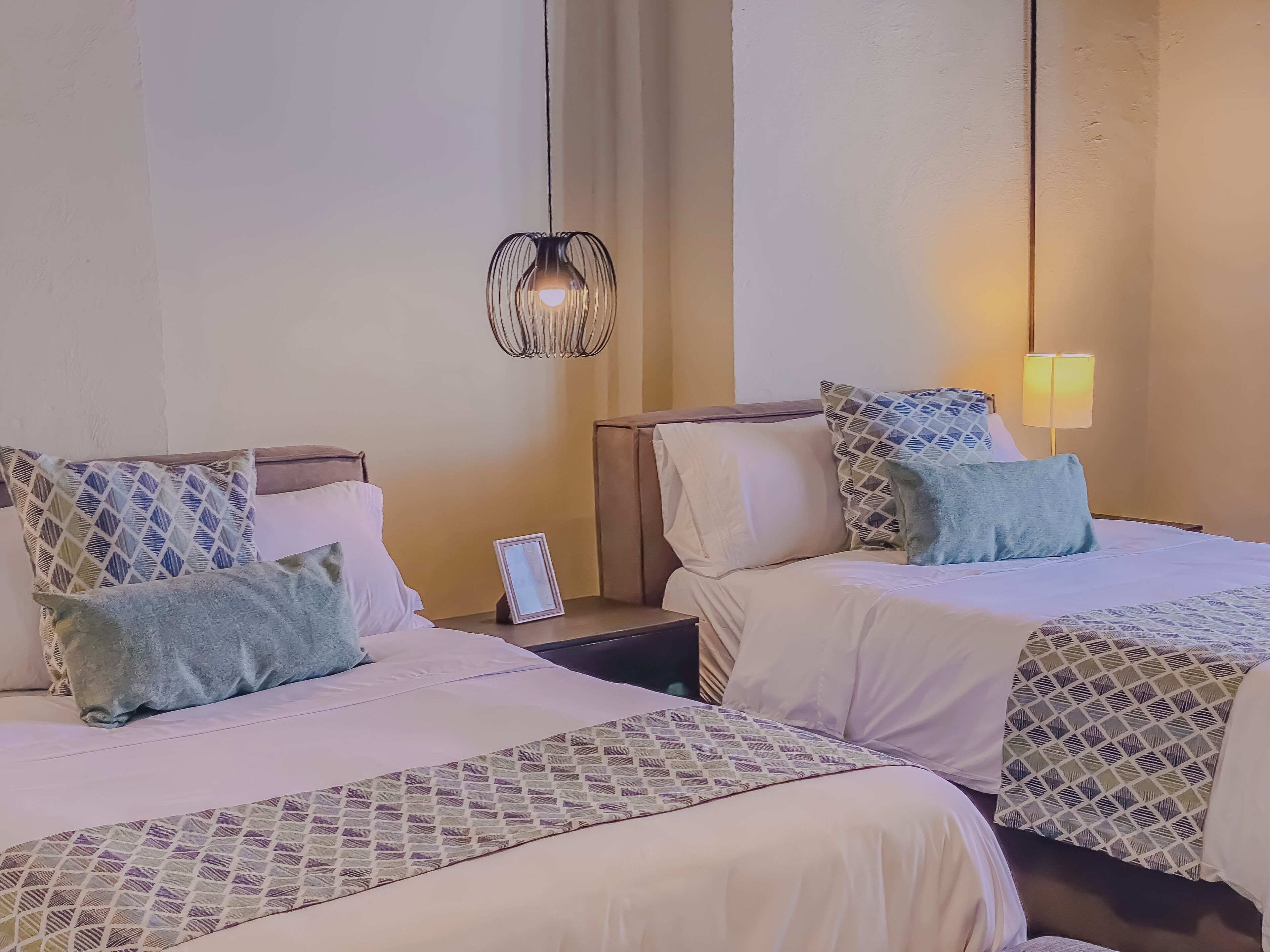 A hotel room with two beds, each with patterned bedding and pillows, a bedside table with a lamp, and a wall-mounted light fixture.