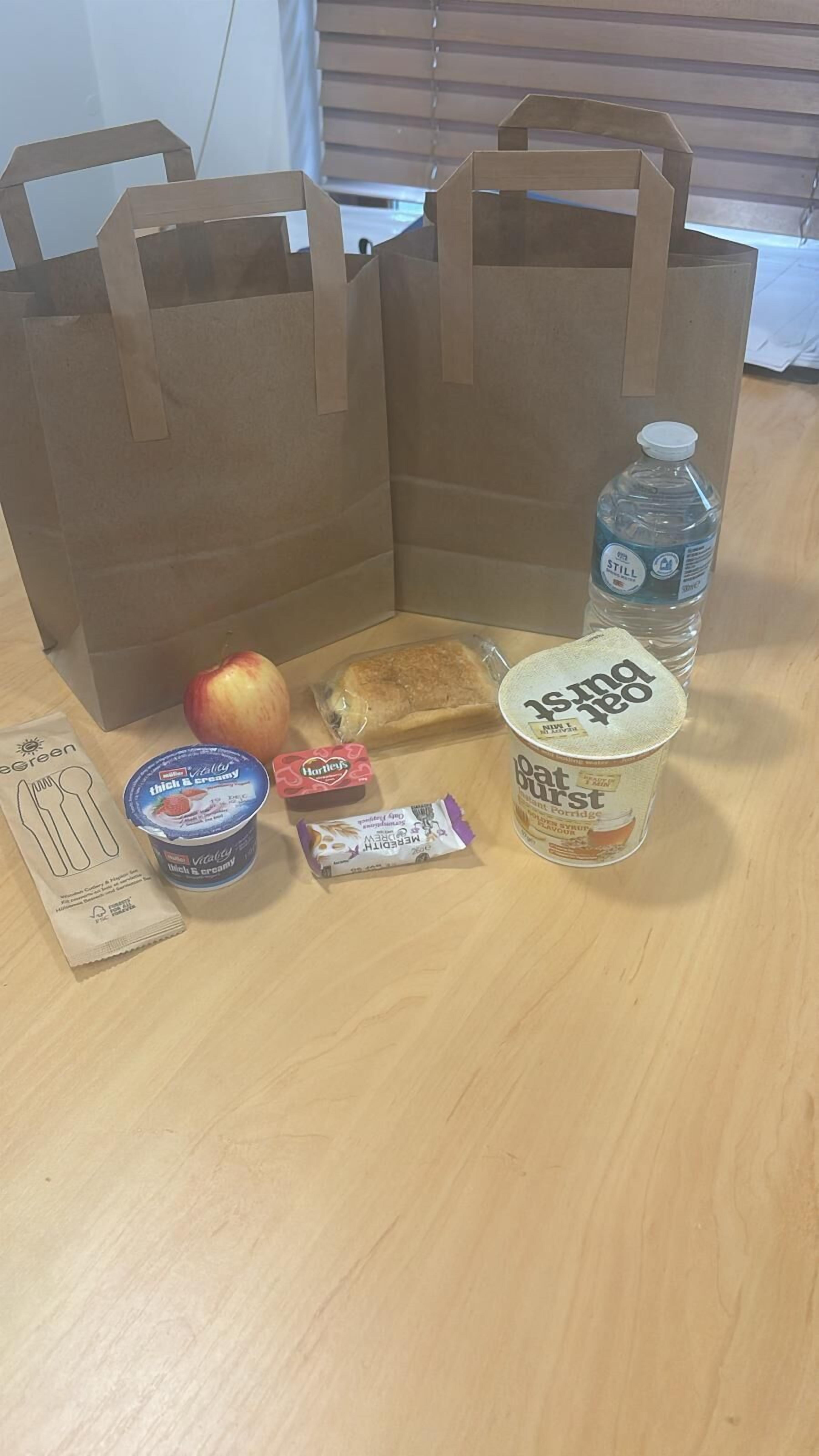 Een houten tafel met twee bruine papieren zakken, een waterfles, een pot met haverrepen, een bakje yoghurt, een pak Hartley’s, een appel in cadeaupapier en een papiertje met tekeningen van bestek.