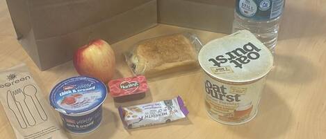 A wooden table with two brown paper bags, a water bottle, a jar of oat bars, a container of yogurt, a packet of Hartley's, a wrapped apple, and a paper with cutlery drawings.
