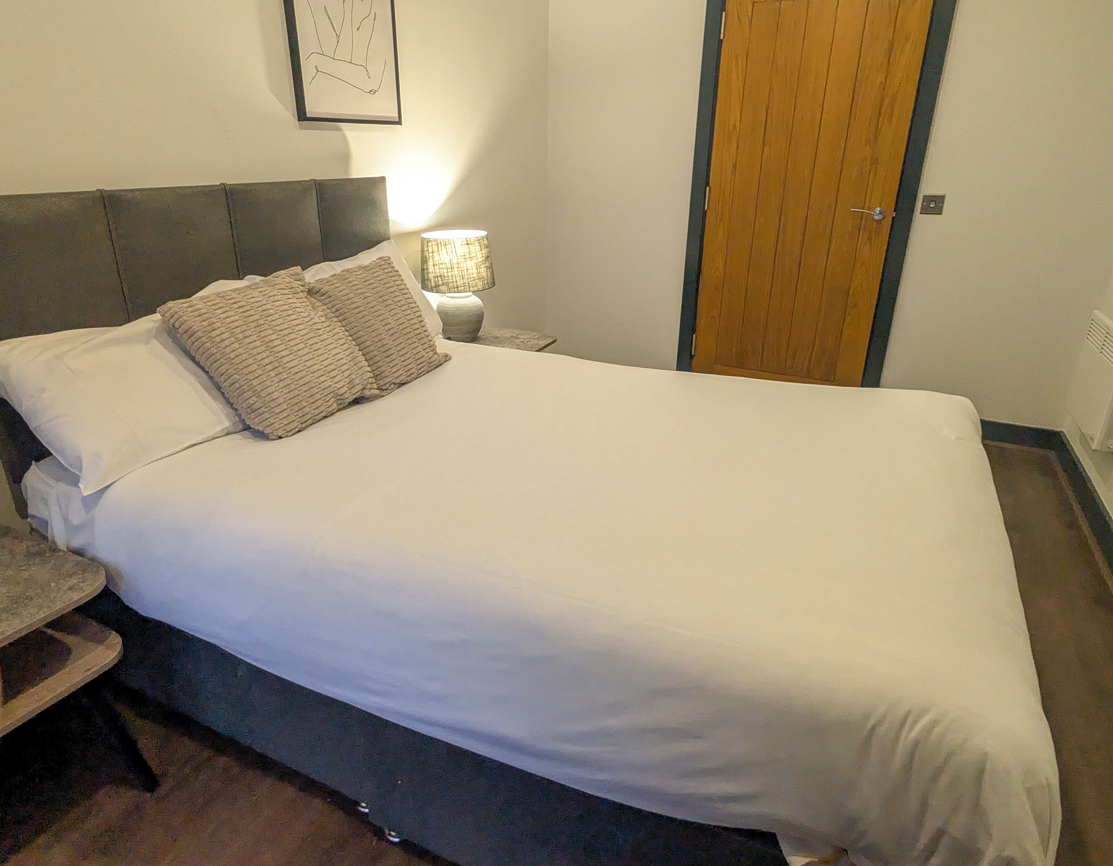 A neatly made bed with white linens and two pillows, a bedside table with a lamp, and a wooden door.