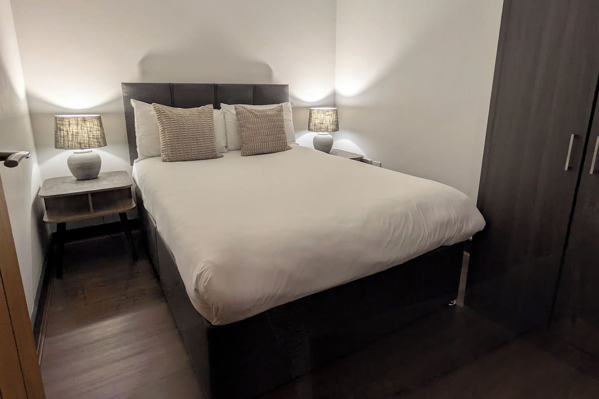 A neatly made bed with a dark headboard, two bedside tables with lamps, and a wardrobe to the right.