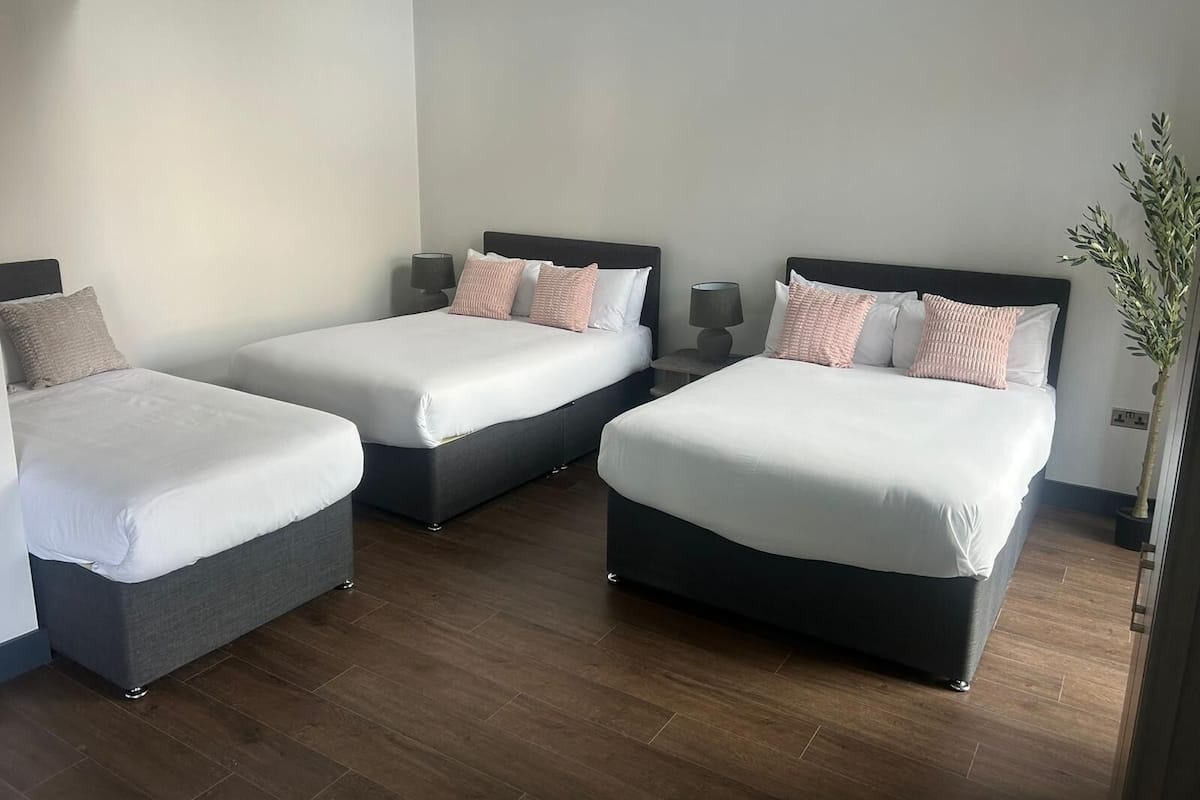 A hotel room with two beds, each with white linens and multiple pillows. There are bedside tables with lamps, a potted plant, and a wooden floor.