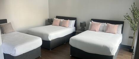 A hotel room with two beds, each with white linens and multiple pillows. There are bedside tables with lamps, a potted plant, and a wooden floor.
