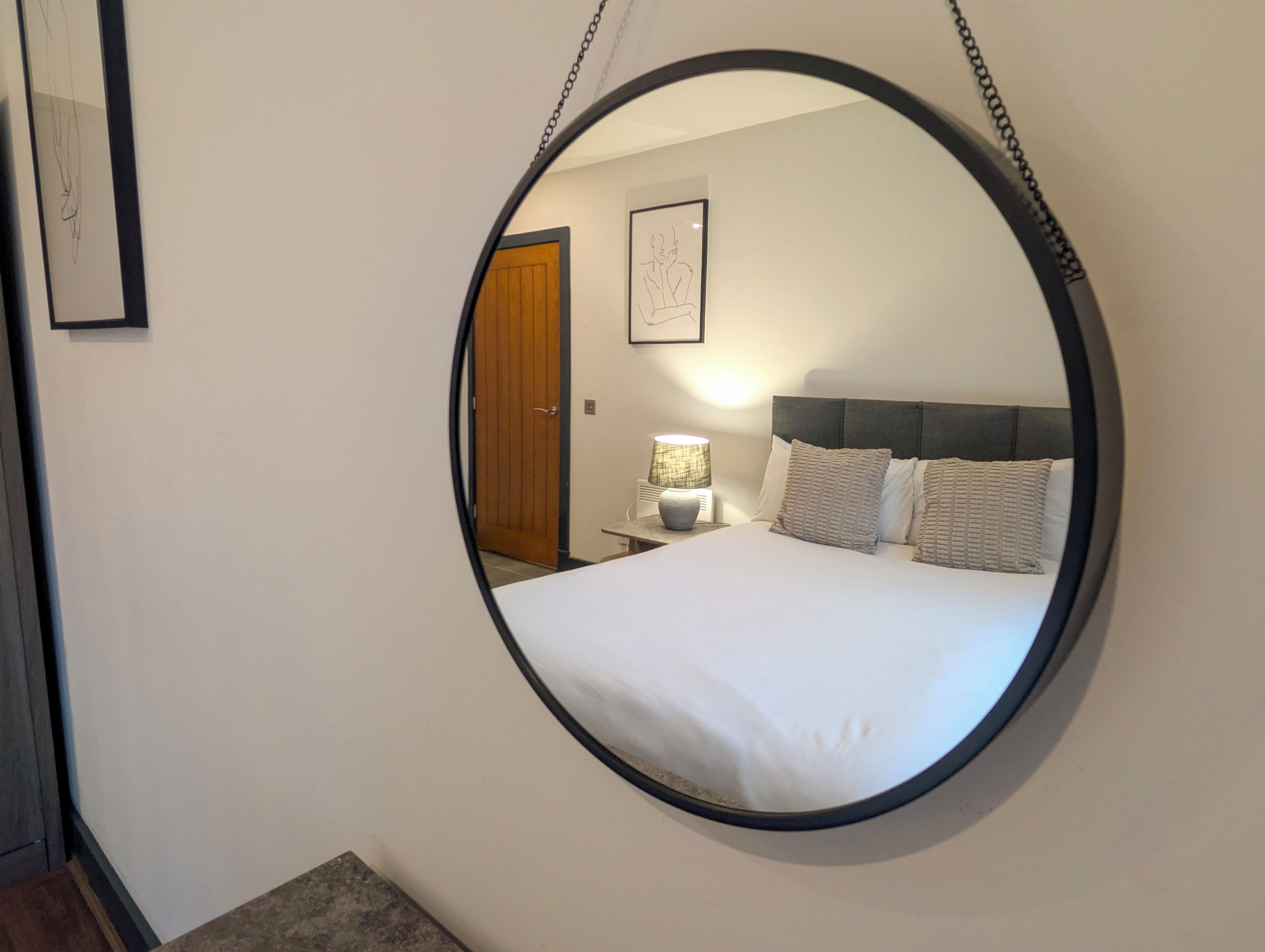 Ein Schlafzimmer mit einem großen, ovalen Spiegel, einem Bett mit weißer Bettwäsche, einem Nachttisch mit Lampe und einer Holztür.