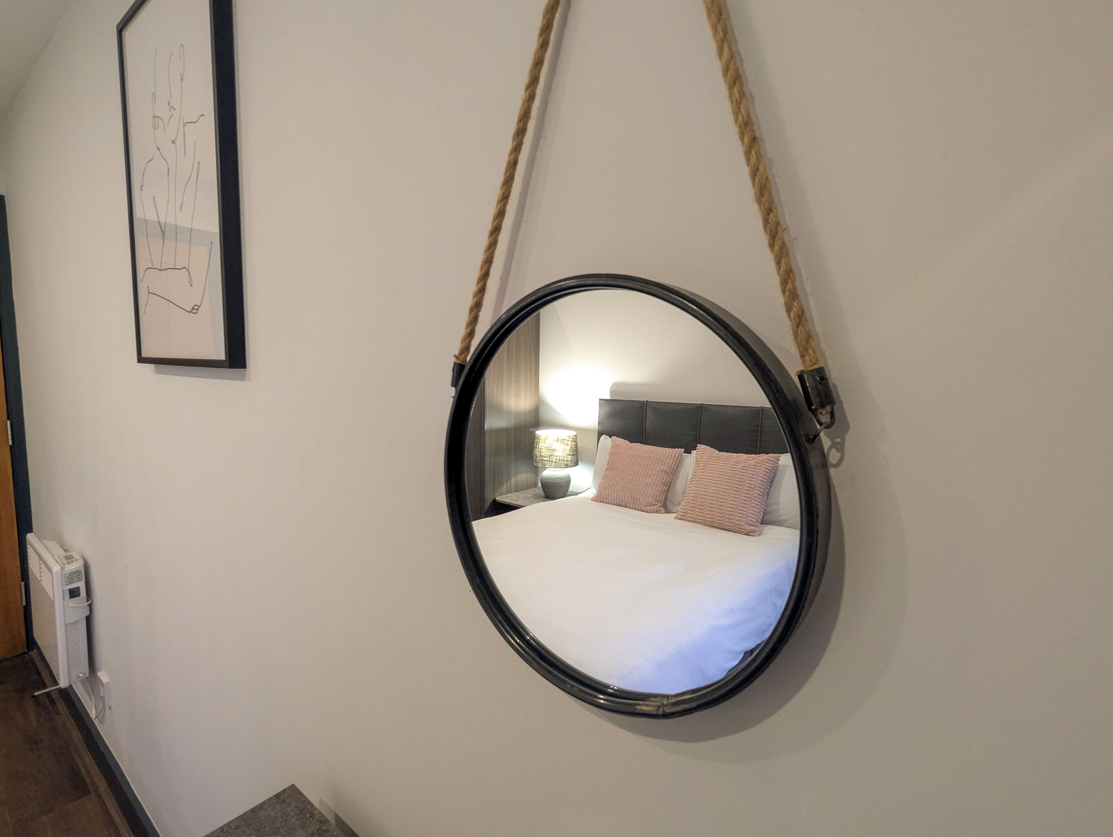 Un miroir rond avec une anse en corde, accroché à un mur blanc, reflète un lit avec des oreillers et une table de chevet sur laquelle se trouve une lampe.