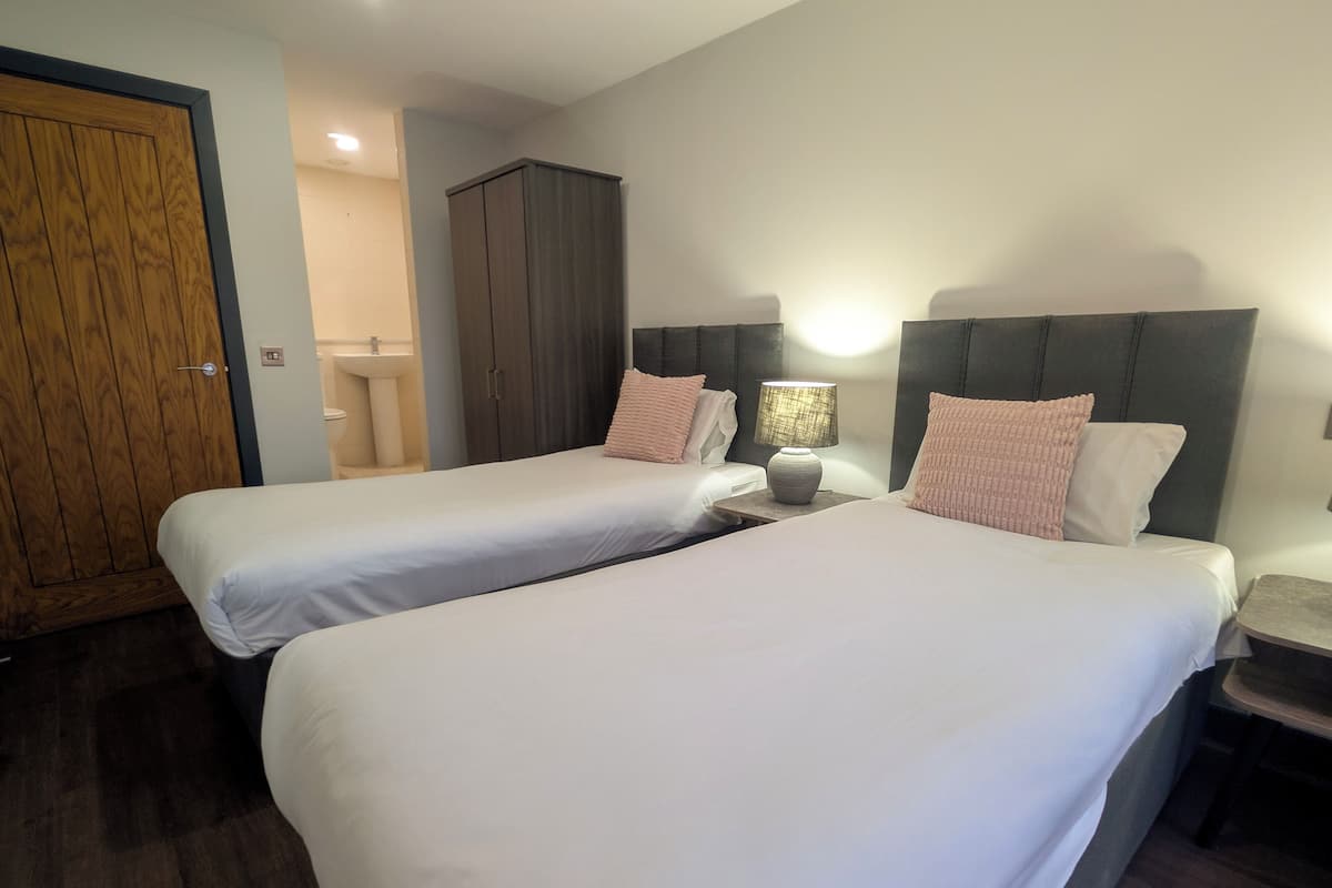 A hotel room with two beds, a wooden wardrobe, a nightstand with a lamp, and a bathroom visible in the background.