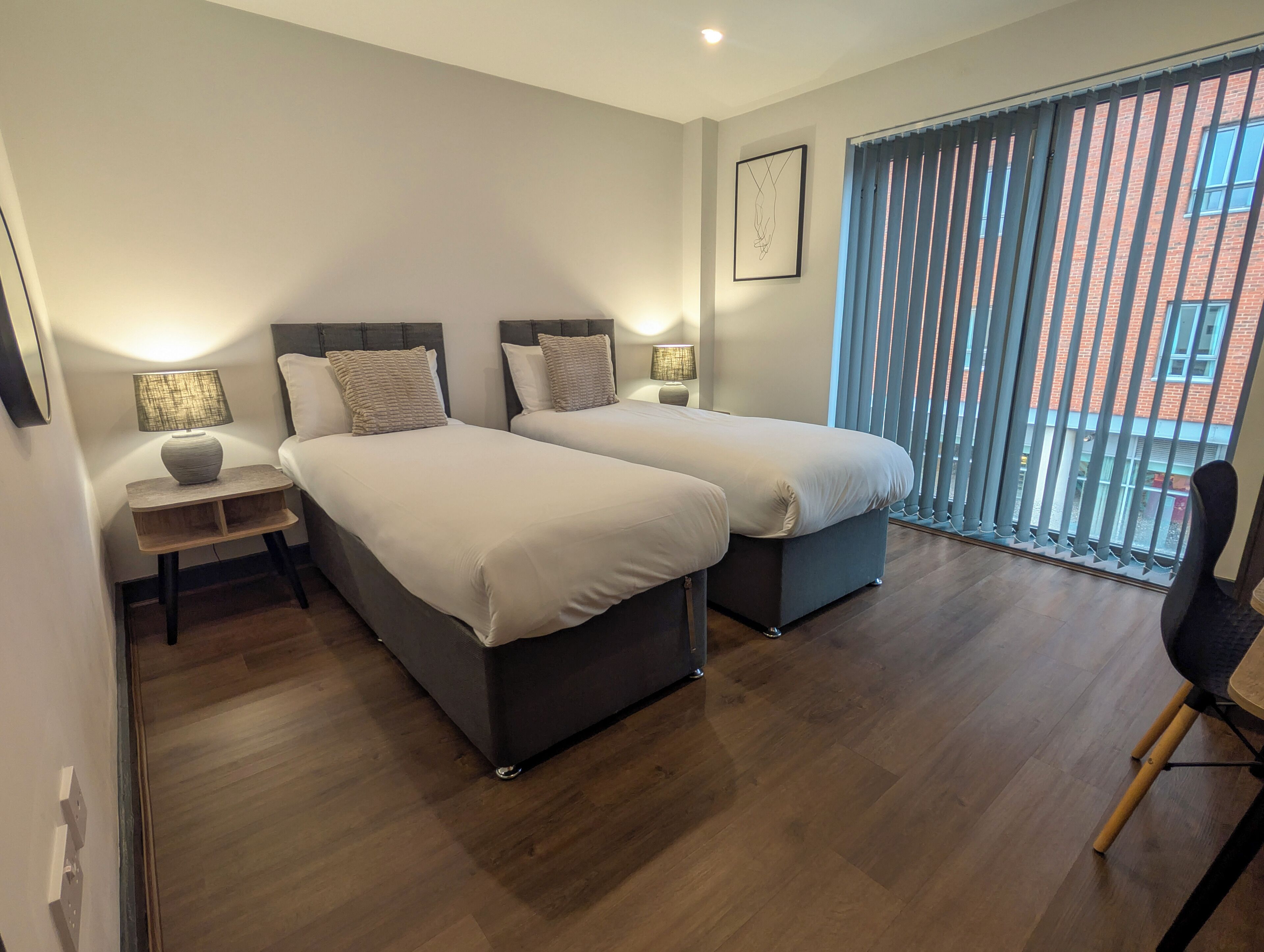 Une chambre moderne avec deux lits, une table de chevet, une chaise et une grande fenêtre avec des stores.