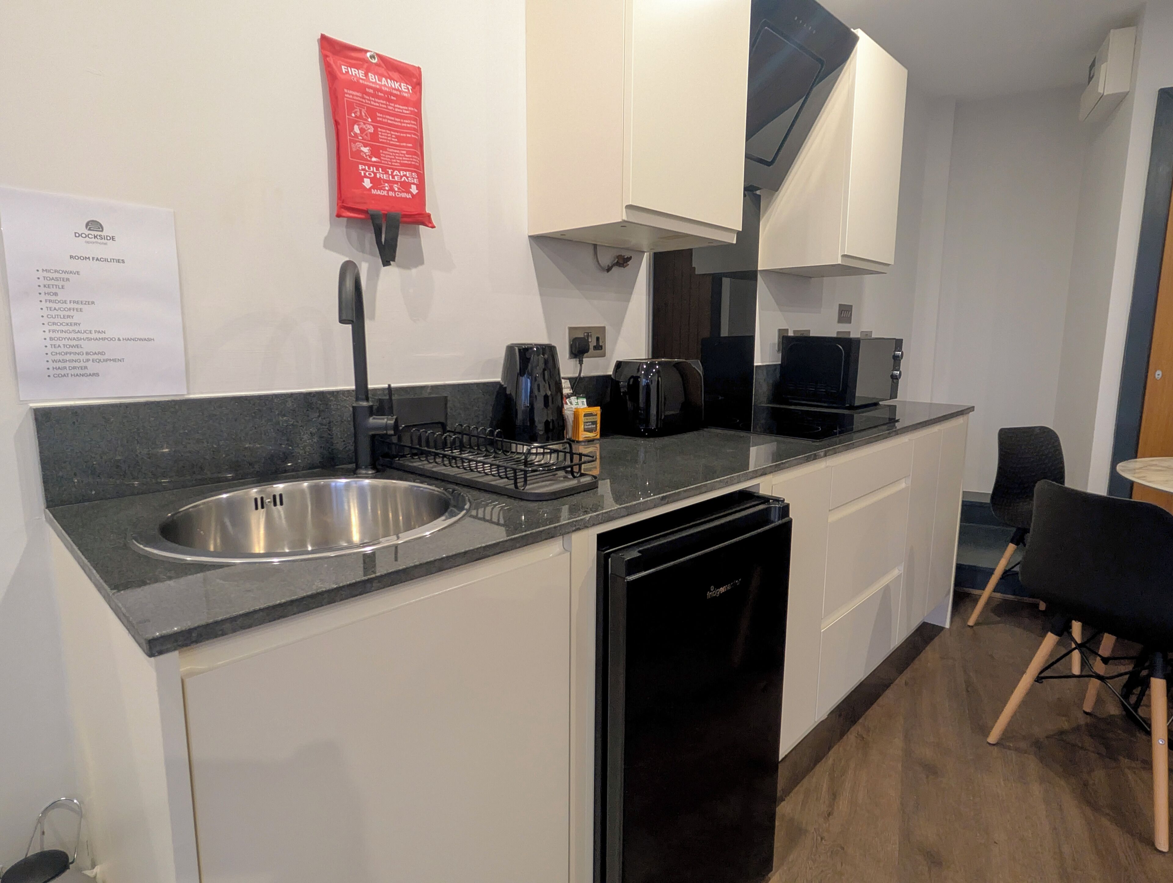 A modern kitchen with a stainless steel sink, black dishwasher, and a coffee maker. A fire blanket is mounted on the wall, and a sign lists room amenities including a microwave and toaster.