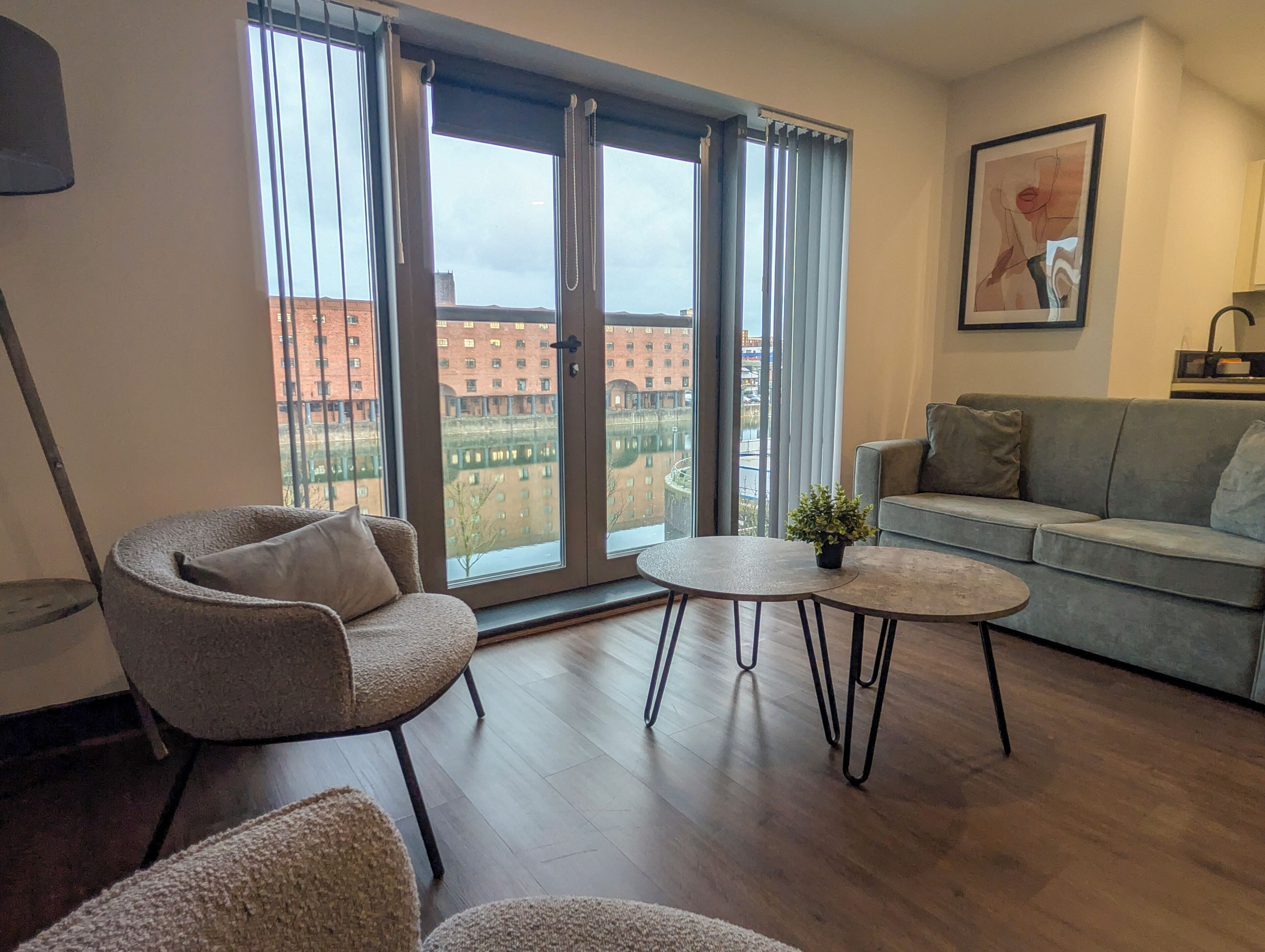 A modern living room with a grey sofa, a round coffee table, and a chair. Large windows offer a view of buildings outside.
