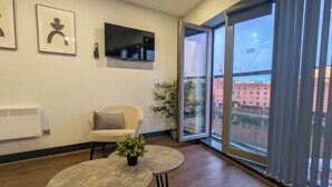 Ein modernes Zimmer mit großem Fenster, einem Flachbildfernseher, einem Sessel, einem kleinen Tisch mit Pflanze und gerahmten Kunstwerken an der Wand.