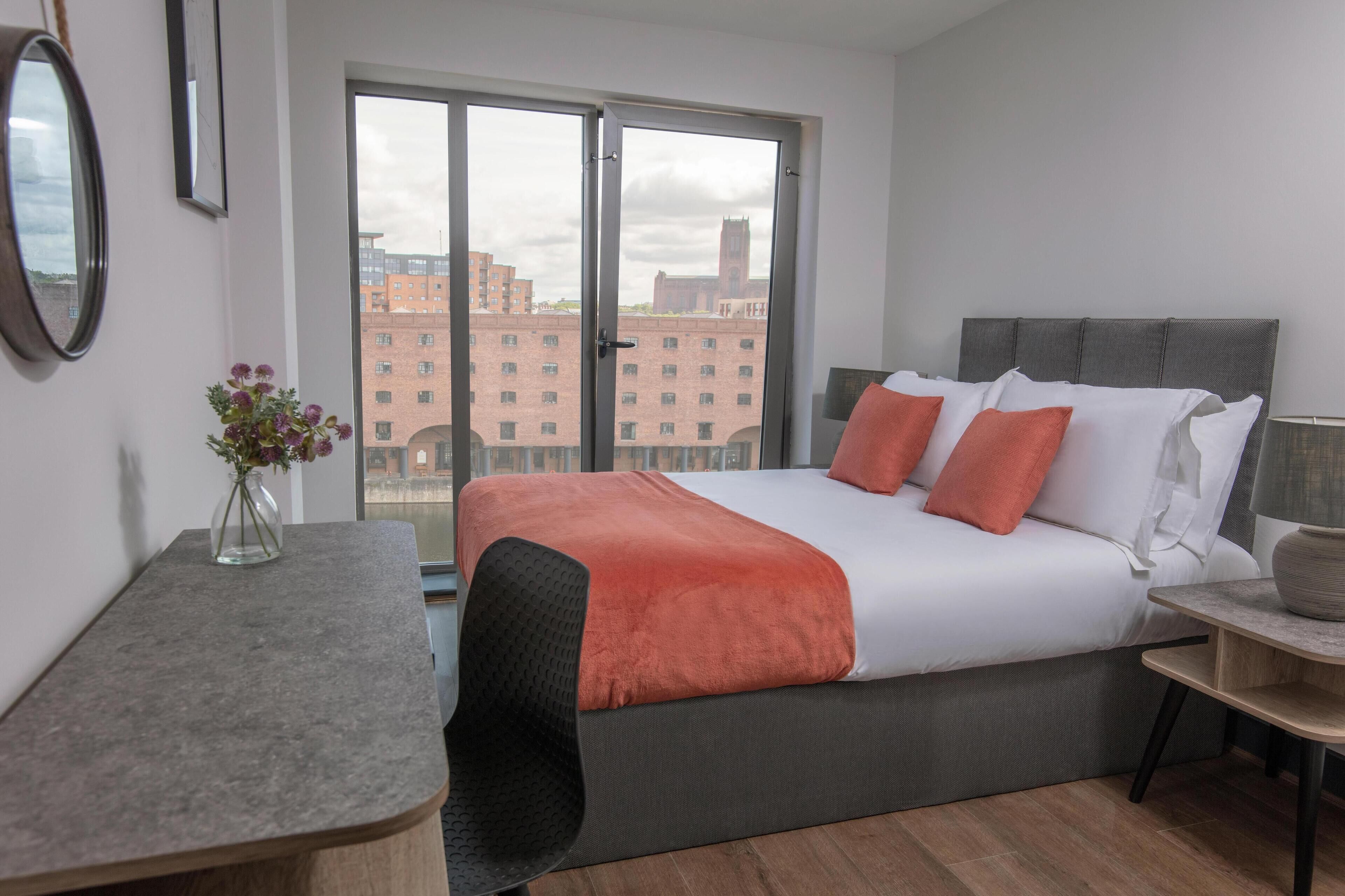 A modern hotel room with a large bed, a desk, and a view of buildings through the window.