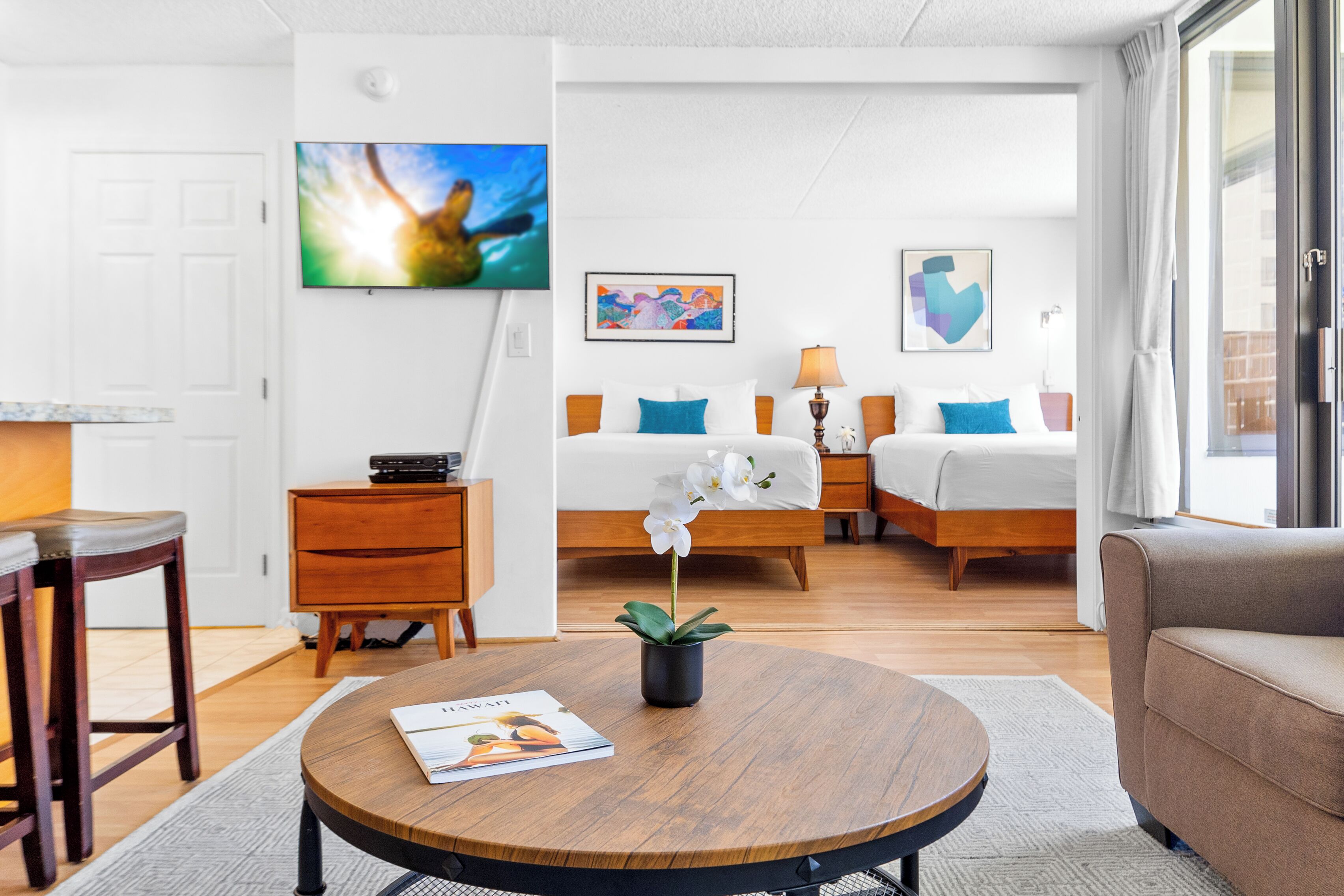 Ett modernt hotellrum med ett soffbord i trä, en säng med blå kuddar, en soffa och en tv som är monterad på väggen.
