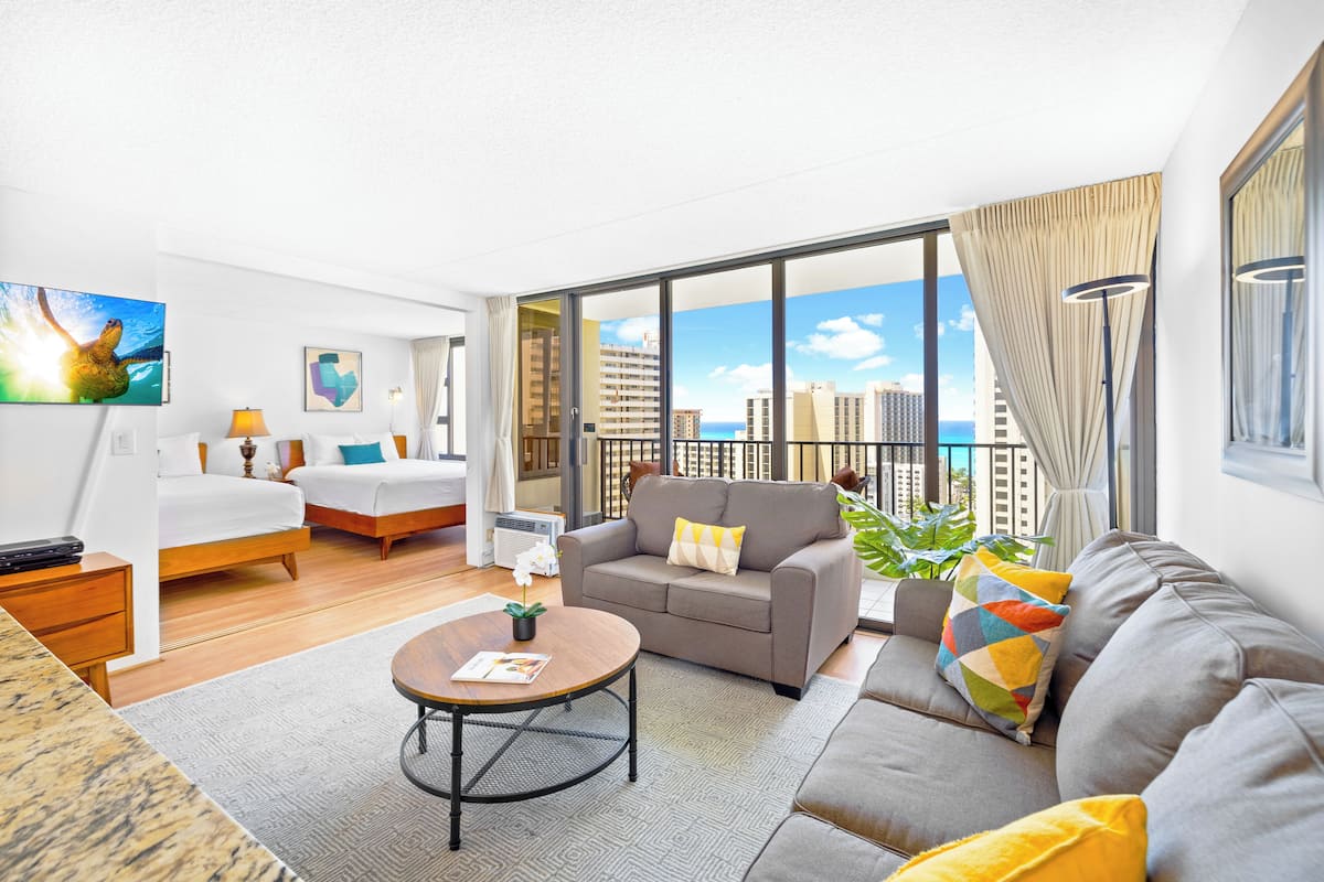 Une chambre d’hôtel moderne équipée d’un lit, d’un canapé et d’une table à manger. Une grande fenêtre offre une vue sur la ville, et une chambre donne sur un balcon.