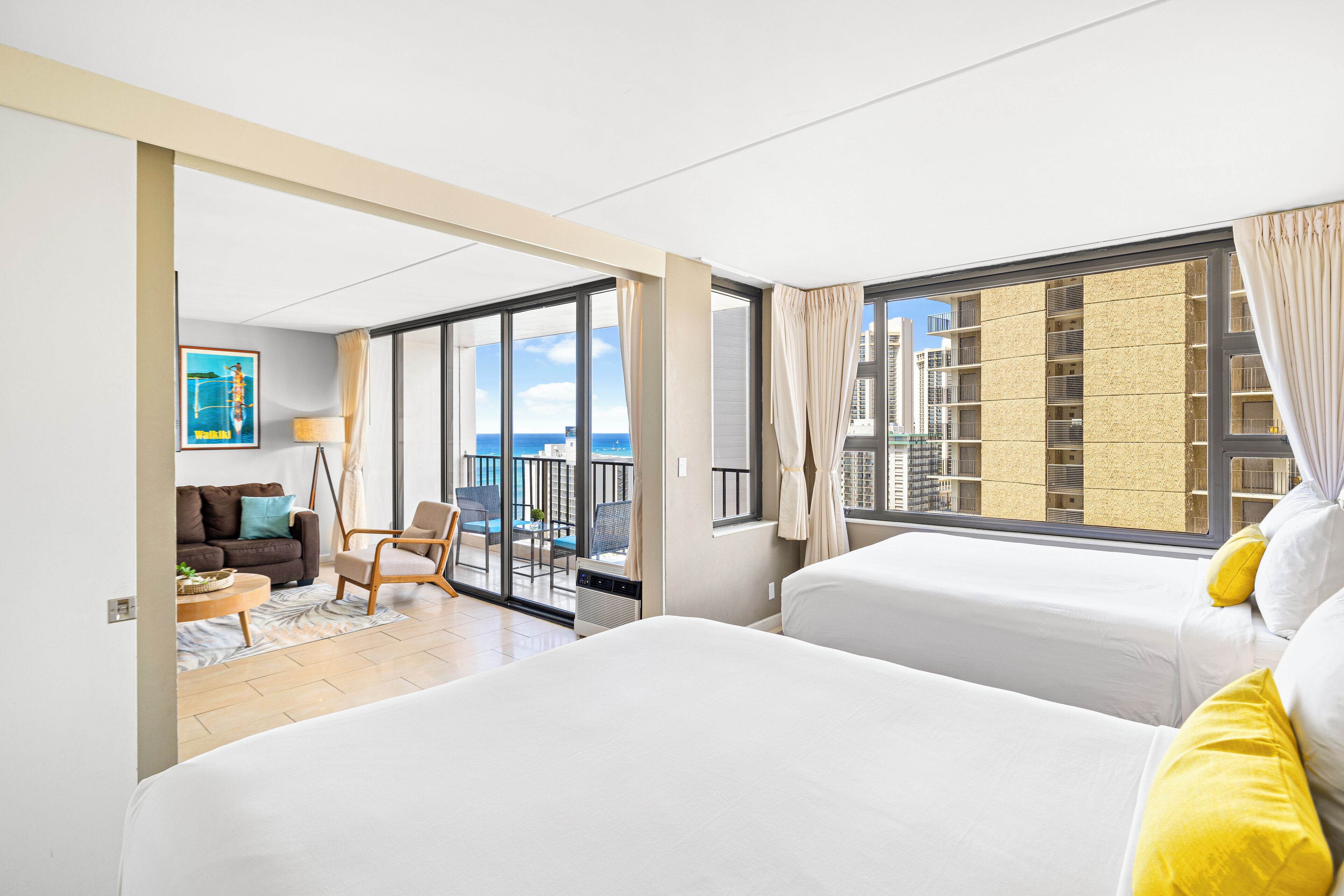A hotel room with two beds, a balcony with a view of the sea, and a dining area with a sofa and chairs.
