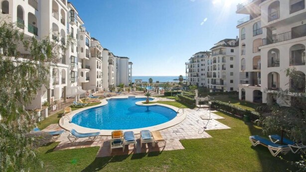 A swimming pool surrounded by a well-maintained lawn and lounge chairs, with multi-story buildings in the background. - Cozy Seaside Apartment in Manilva (Manilva)