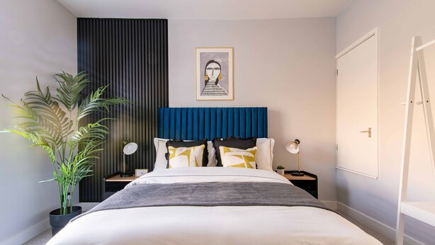 Ein modernes Schlafzimmer mit einem großen Bett, einem blauen, gepolsterten Kopfteil, einem Blumentopf, einem gerahmten Kunstwerk an der Wand und einer weißen Tür.