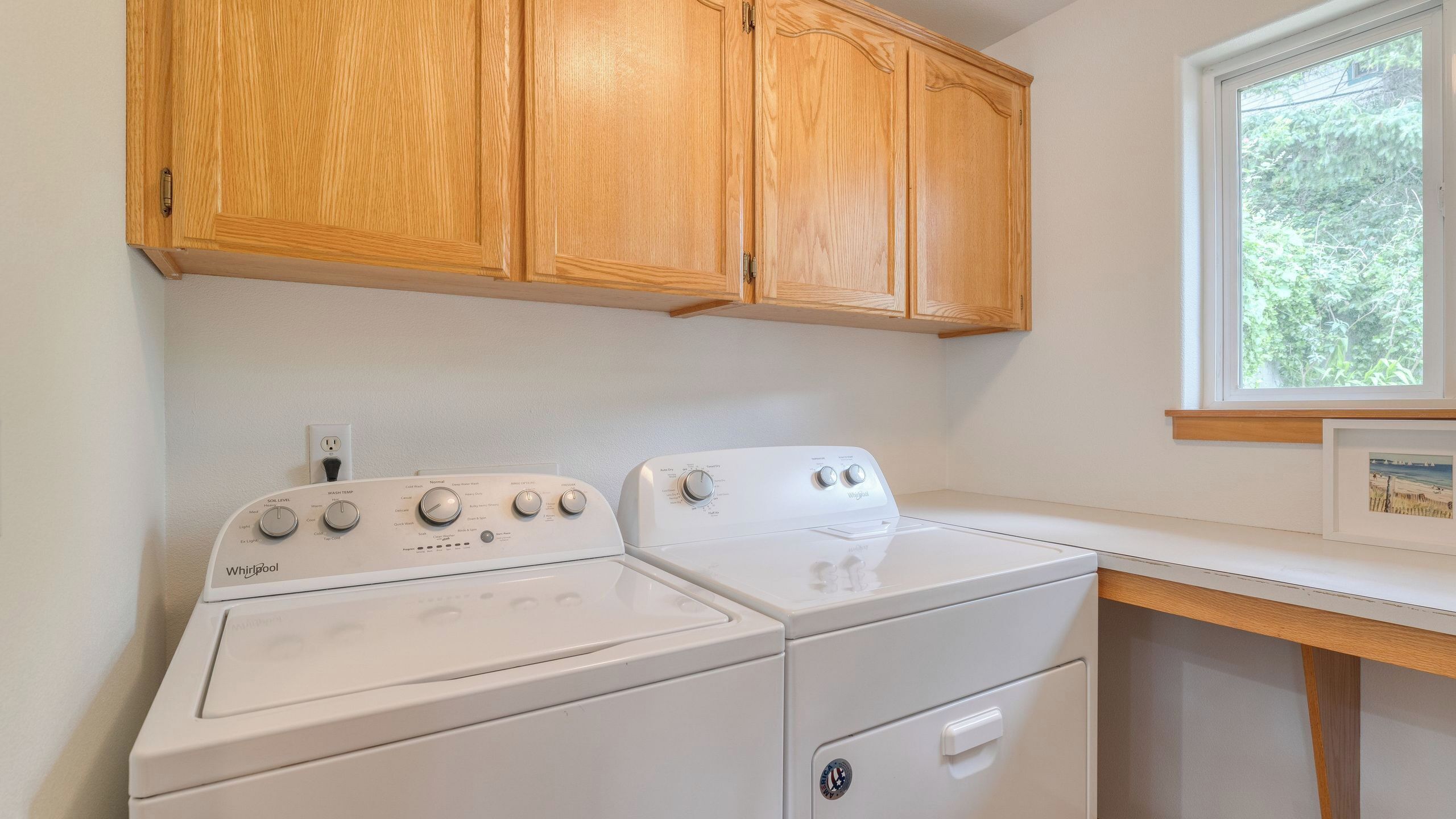 A laundry room with two Whirlpool washers, wooden cabinets, a window, and a countertop.