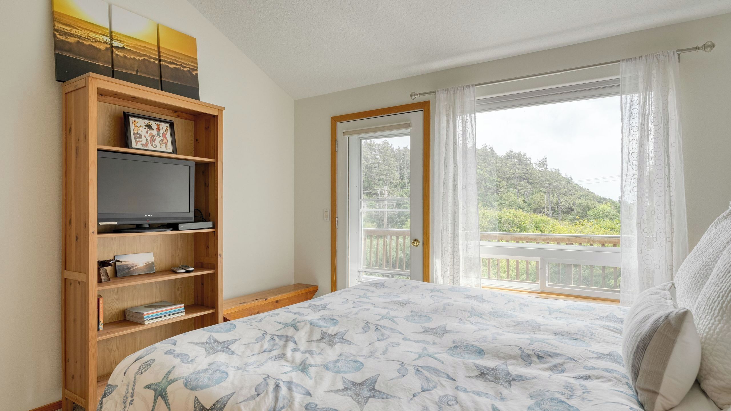 Een slaapkamer met een bed, een televisie op een houten plank, een raam met uitzicht op groen, en een deur.