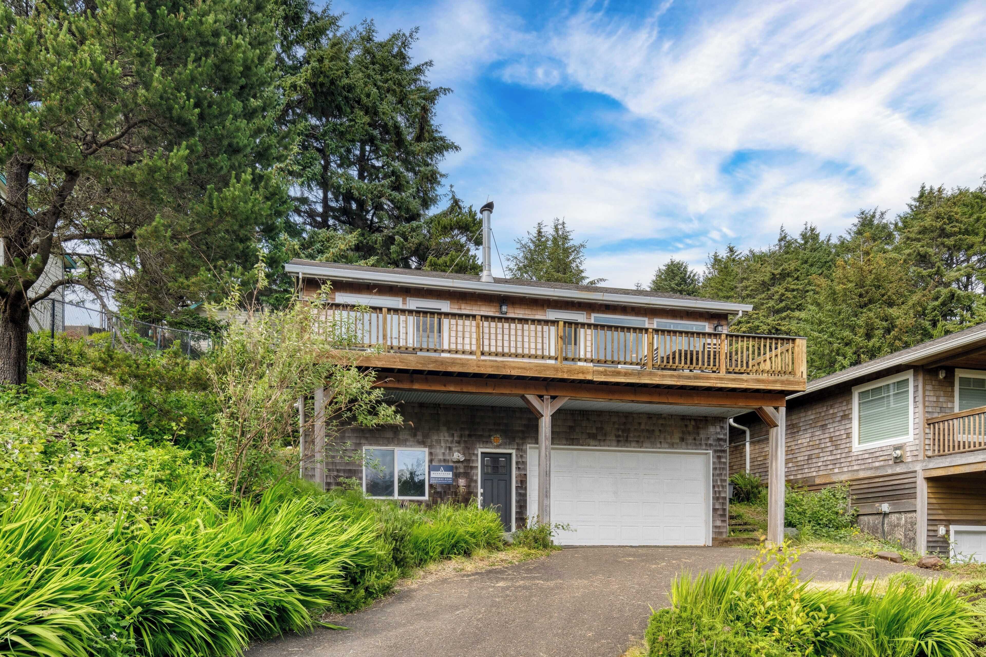 A two-story house with a balcony, a garage, and a driveway surrounded by greenery.